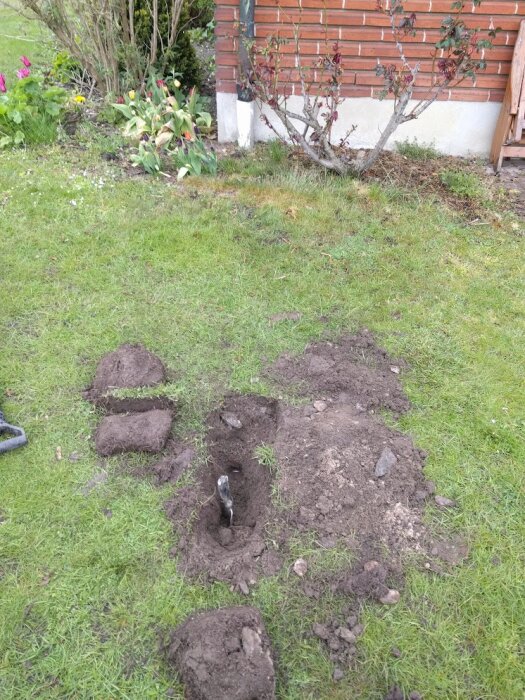 Grävd grop i en trädgård bredvid en tegelvägg, med uppgrävd jord och bitar av grästorv runtom. En spade sticker upp från hålet.