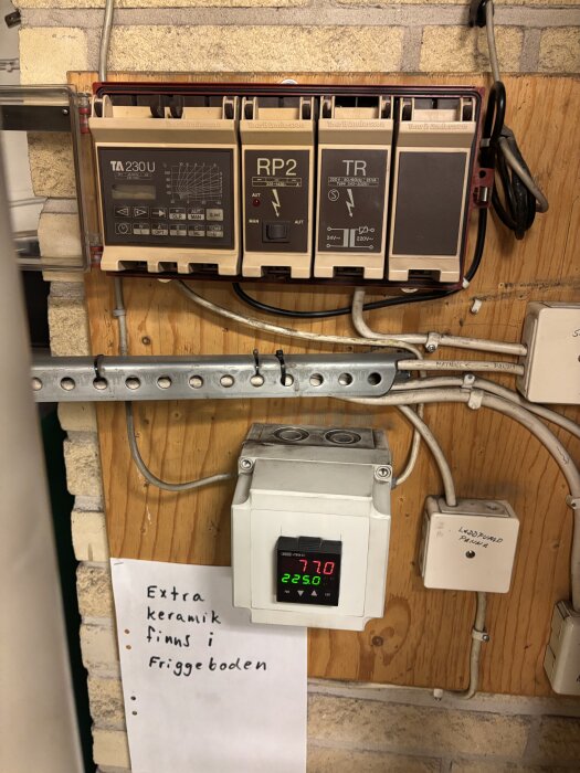 Gamla elektriska regulatorer och en termostat är monterade på en träpanel. Text noterar "Extra keramik finns i friggeboden".