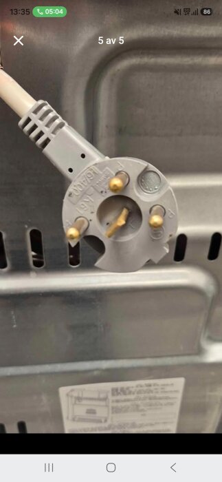 Grå apparatkontakt med tre stift, använd för anslutning till ett 19V uttag, mot en metallbakgrund av en hushållsapparat.