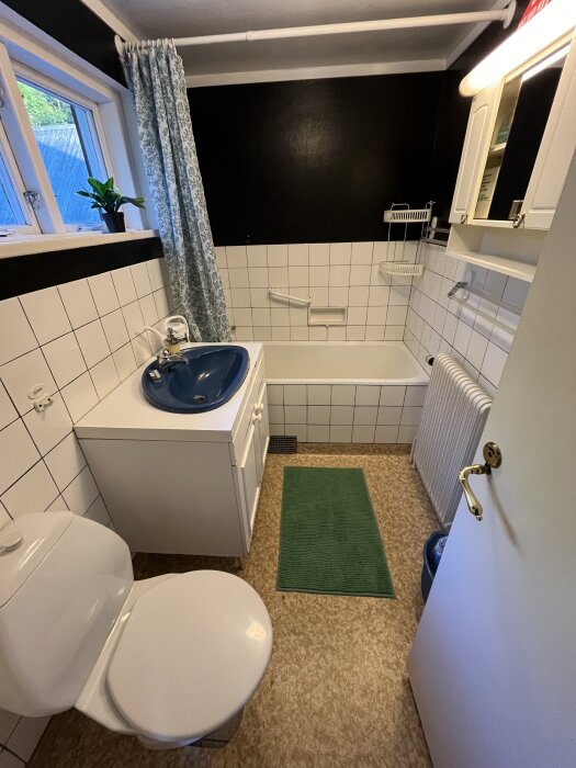 Badrum från 1960-talet med vit kakel, badkar, blå tvättställ, toalettstol och radiator. Grön matta på golvet och duschdraperi vid fönstret.