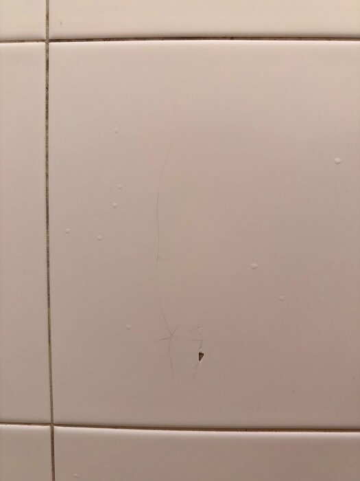 Sprucket vitt kakel i dusch med synliga sprickor och liten skada som exponerar underlaget.
