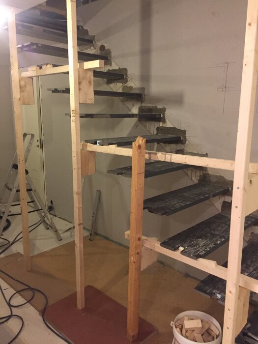 Trappa under byggnation med ställning av trä runt ståltrappsteg fästa i en betongvägg, visar stegkonstruktion och förankring i väggen.