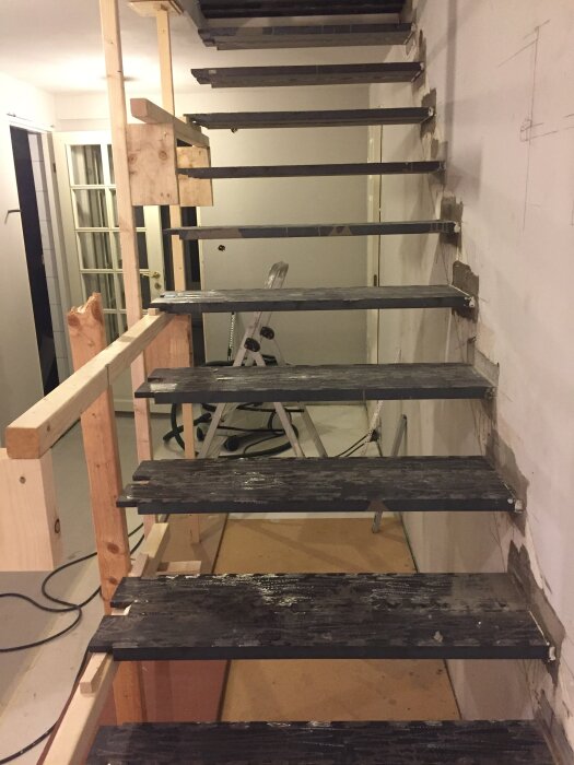 Trappa under byggnation med kraftiga stålplåtsteg, monterade i en Leca-vägg. Träställningar hjälper med installationen. Bakgrund av oavslutad inomhusrenovering.