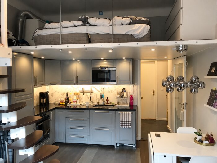 Funktionellt kök under loft med gråa skåp, marmorkakel, spiraltrappa till loft, modern belysning, matbord och loftsäng i ett kompakt boende.