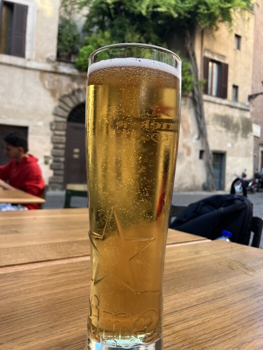 Ett glas öl på ett träbord utomhus vid en restaurang, med suddig stadsmiljö och vegetationen i bakgrunden.