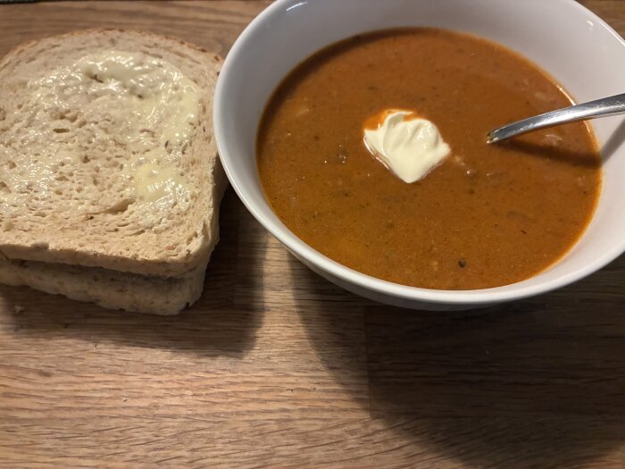 En skål med gulasch med gräddklick, serverad med två brödskivor med smör, på ett träbord.