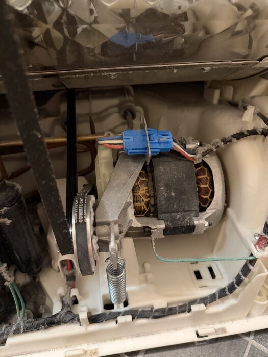 Interiör av en tvättmaskinmotor med rem och fjäder, visar kablar och komponenter i maskinens mekaniska del.