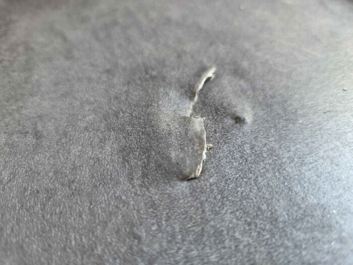 Skada i våtrumsmatta med en ca 5 cm lång reva och en buckla, orsakat av tappad värmepump, synlig innanför dörren.