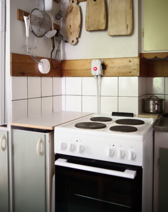 1960-talskök med ny elspis, vita plattor, träpanel, säkerhetsbrytare med röd signallampa, temperaturvakt och hängande köksredskap.