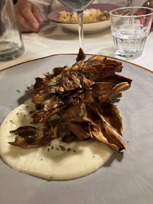 Friterad kronärtskocka på en bädd av krämig sås, garnerad med gröna örter, serverad på en tallrik med andra rätter i bakgrunden.