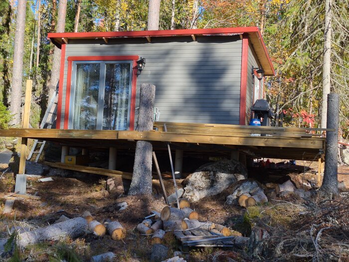 Litet fritidshus i skogen, med grå fasad och rött tak, delvis färdig altan och trädstammar i förgrunden.