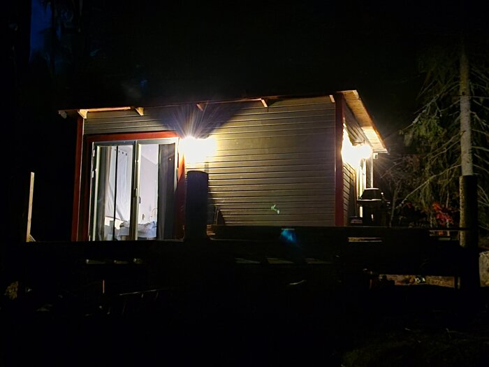 Upplyst stuga i skogen med en glasdörr och inre glimt av ett sovrum, nattmiljö med träd i bakgrunden.