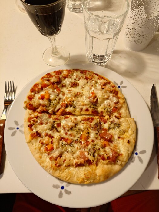 Pizza med skinka, tomat, lök och vitlök på en vit tallrik med ett glas rött vin och ett glas vatten bredvid.