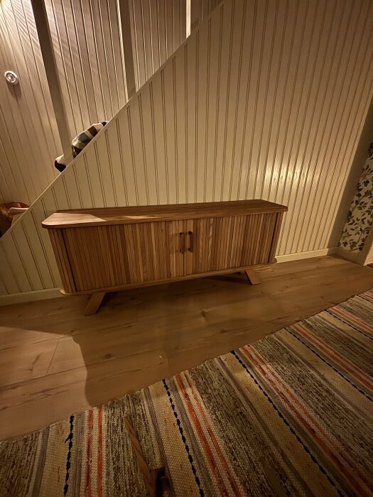 Träskåp med ribbad design står mot vit panelvägg, placerad på ett trägolv bredvid en randig matta.