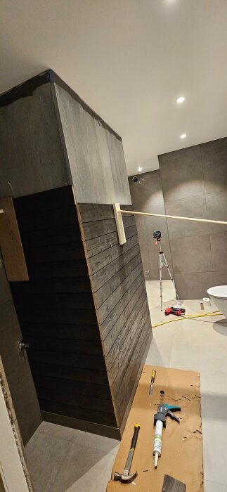 Badrum med pågående renovering, en murstock klädd i mörkt träpanel, verktyg och kartong syns på golvet.