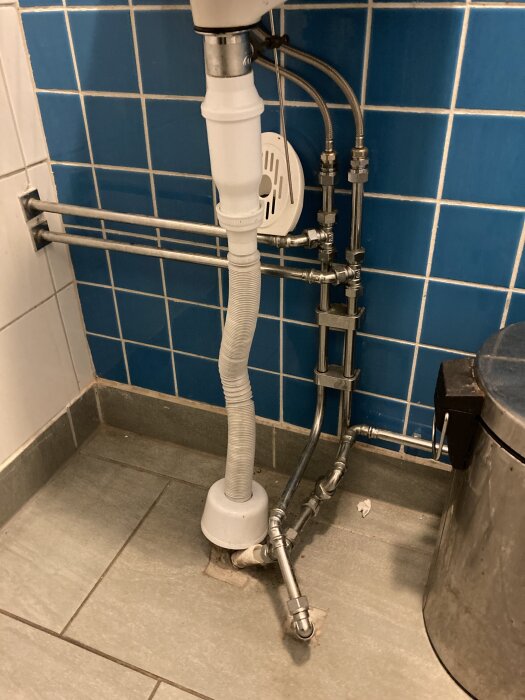 Rörledningar och vattenlås under ett tvättställ i ett badrum med blå kakelvägg och grått klinkergolv.