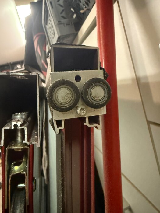 Närbild på skjutdörrskena med två hjul monterade, del av en mekanism för skjutdörrar mot en bakgrund med bland annat en röd stång.