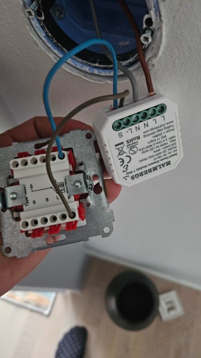 En hand håller i en elektrisk dosa med flerfärgade kablar som är delvis anslutna. Bakgrunden visar en vägg och golv.