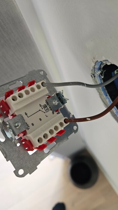 Elektrisk kopplingsdosa med grå och brun kabel monterad, synlig vägguttag i bakgrunden.