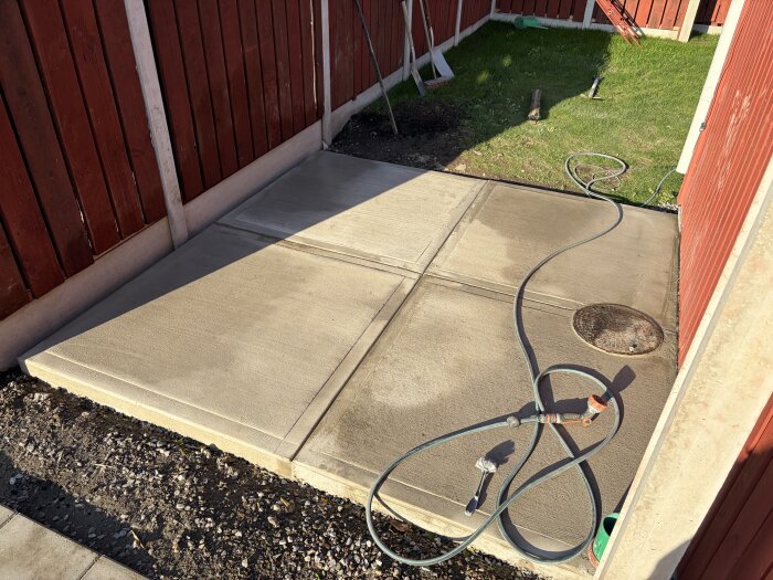 Betongplattor i en hörna av en trädgård med röd trästaket, vattenslang och borste på ytan.