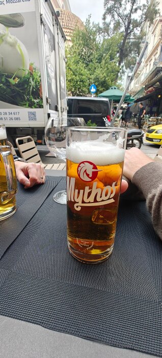 Ett glas Mythos-öl står på ett bord utomhus, omgiven av människor och trafik på en livlig gata.