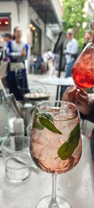 Ett glas rosa cocktail med is och gröna blad, stående på ett bord utomhus vid en trottoar med suddiga människor i bakgrunden.