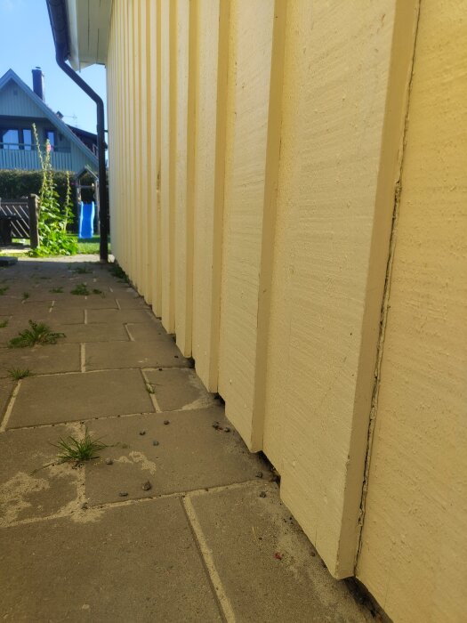Gulmålad husfasad med träpanel och glipor längst ner, fotograferad från sidan på en stengång, med andra hus och trädgårdar i bakgrunden.