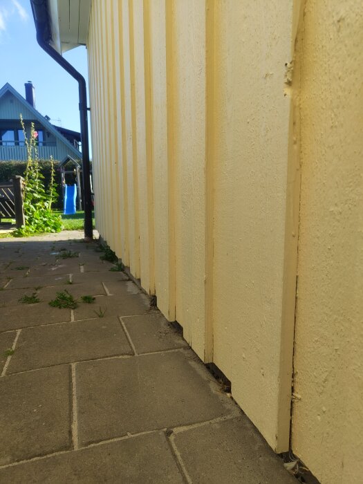 Gulmålad husfasad med glipor vid botten, ligger intill stenlagd gång med ogräs; visar behov av förbättrad tätning och ventilation innan ommålning.