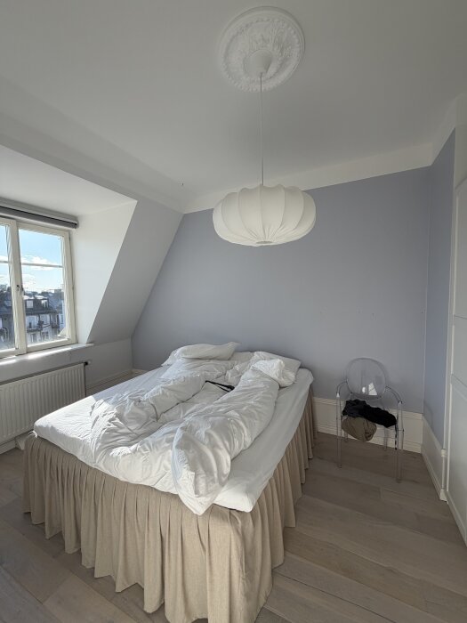 Sovrum med en dubbelsäng, vita sängkläder, ljust trägolv, plaststol och frodig taklampa. Blå vägg med snedtak och fönster.