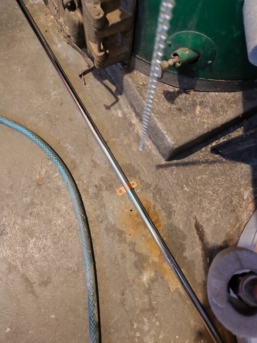 Metallstång med fäste på betonggolv bredvid en grön stor tank och en blå vattenslang.