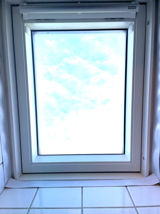 Velux takfönster med ljusinsläpp, omgiven av vita träkanter och kakel. Tejp rester synliga längs kanterna. Kortsidorna ser olika ut jämfört med långsidorna.
