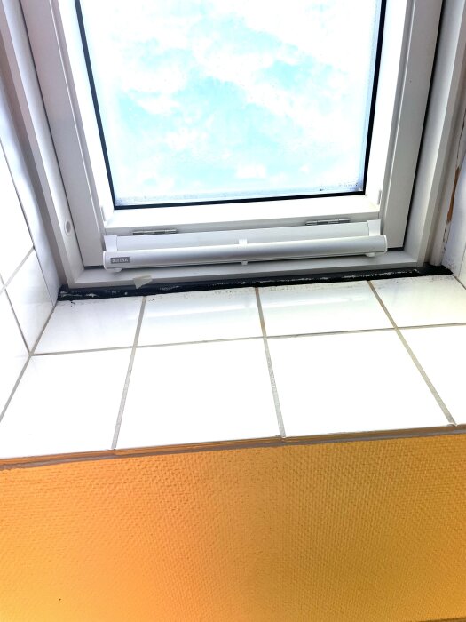 Veluxfönster installerat med vitt plastram, omgiven av kakel och orange vägg. Tejp rester och färgskada synliga på ramen, saknar invändig list.