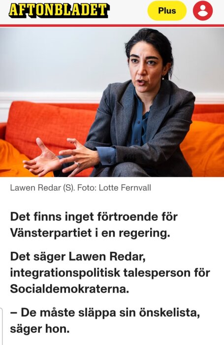 En person i grå kostym diskuterar engagerat i en soffa med orange bakgrund.