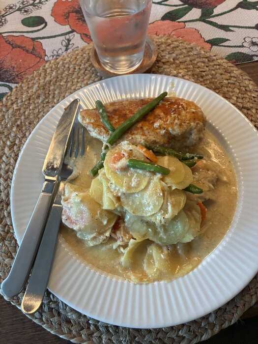 Kycklingfilé och skivad potatis med morot i sås, serverad på en vit tallrik med gröna bönor, bredvid kniv och gaffel.