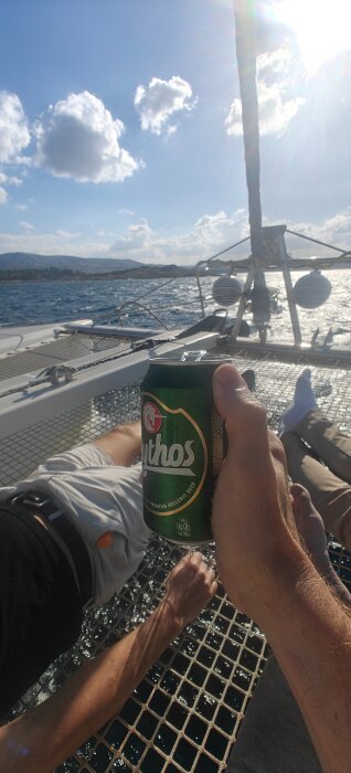 En person håller en ölburk av märket Mythos på en segelbåt med personer som solar på nätet, medan båten glider över havet under en solig dag.