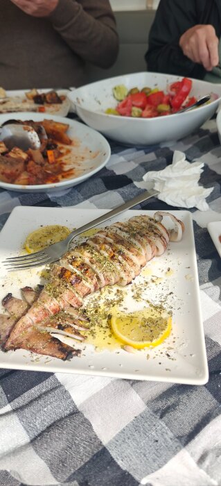 Grillad bläckfisk skuren i skivor med kryddor och citronskivor, uppdukat på ett vitt fat med sallad och grönsaker i bakgrunden.