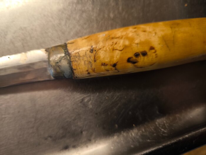 Handtag av masurbjörk med mörka mönster, som associeras till ansikts- och djurmönster, fäst vid en kniv med en metallbrygga.