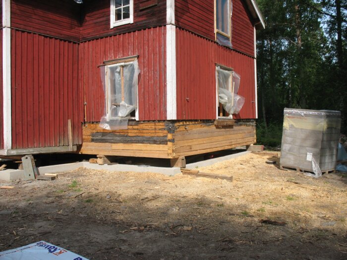Renovering av rött trähus, grundarbete synligt. Fönster skyddade med plast. Byggmaterial i förgrunden. Skog i bakgrunden.
