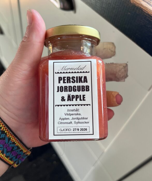 Burk med persika, jordgubb och äppelmarmelad, handhållen med etikett som visar ingredienser och tillverkningsdatum 27/9 2026.