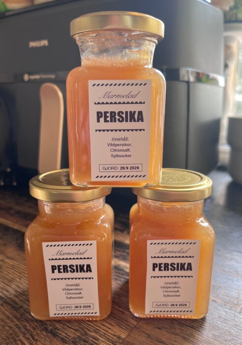 Tre burkar hemgjord persikomarmelad med etiketter som visar ingredienser: vildpersikor, citronsaft, syltsocker. Gjord 26/9 2026.