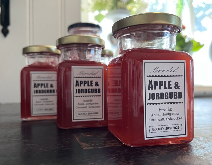 Burkar med äpple- och jordgubbsmarmelad i soligt kök, etiketterade med ingredienser och tillverkningsdatum.