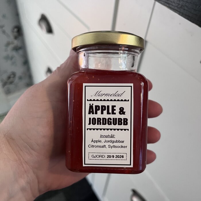Burk med hemgjord äpple- och jordgubbsmarmelad, etikett visar ingredienser och tillverkningsdatum, perfekt för vinterförvaring eller som julklapp.