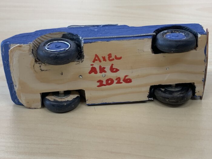 Trämodell av en bil sedd underifrån med hjul och texten "Axel ÅK 6 2026" målad i rött på träets undersida.