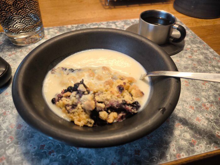 Blåbärspaj med vaniljsås i en svart skål, serverad på ett mönstrat bord med ett glas vatten och en kopp kaffe intill.
