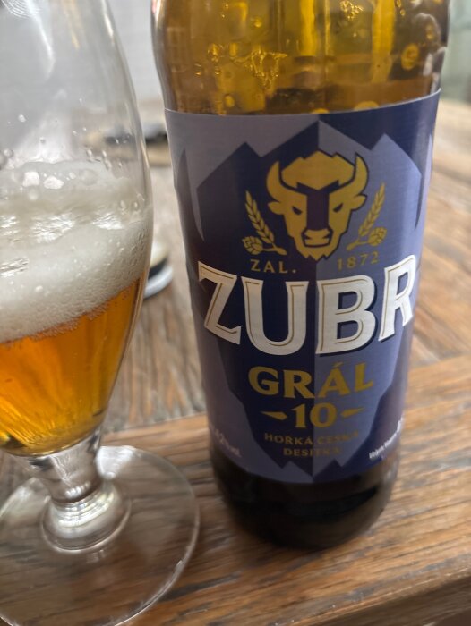 Flaska och glas med Zubr Grál 10 öl på ett träbord. Etiketten visar en buffel och texten "Horká Česká Desítka".