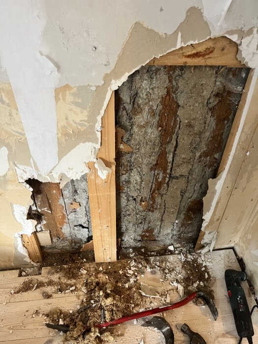 Vägg under renovering med riven tapet och blottlagt trä, gips och lerklining synliga. Verktyg och byggskräp på golvet.