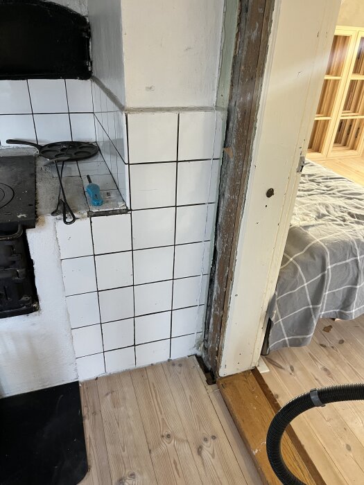 Köksvägg med vit kakel, synlig plankstomme och trägolv. En stekpanna och skruvmejsel ligger på en svart arbetsyta.