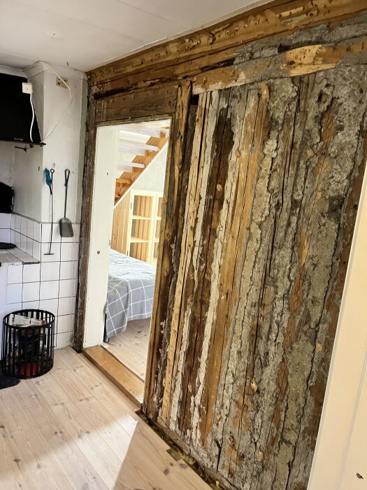 Riven vägg med synlig plankstomme och lerklining bredvid köket, visar en del av ett saneringsprojekt med trasigt ytskikt.