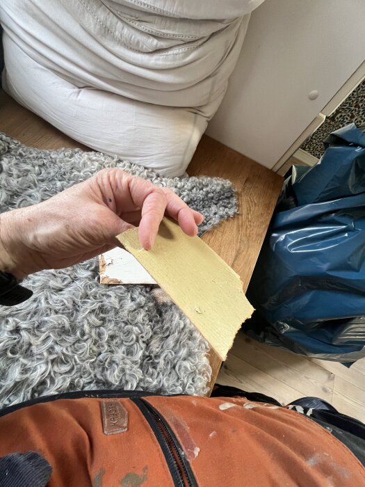 Hand håller gulbrun bit av gammal väggbeklädnad ovanpå grått fårskinn och brunt tyg, på golvet syns även en blå sopsäck.