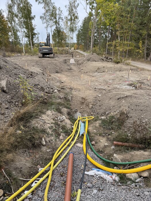 Byggarbetsplats med grävmaskin, installationsrör och fyllnadsmassor; skog i bakgrunden.